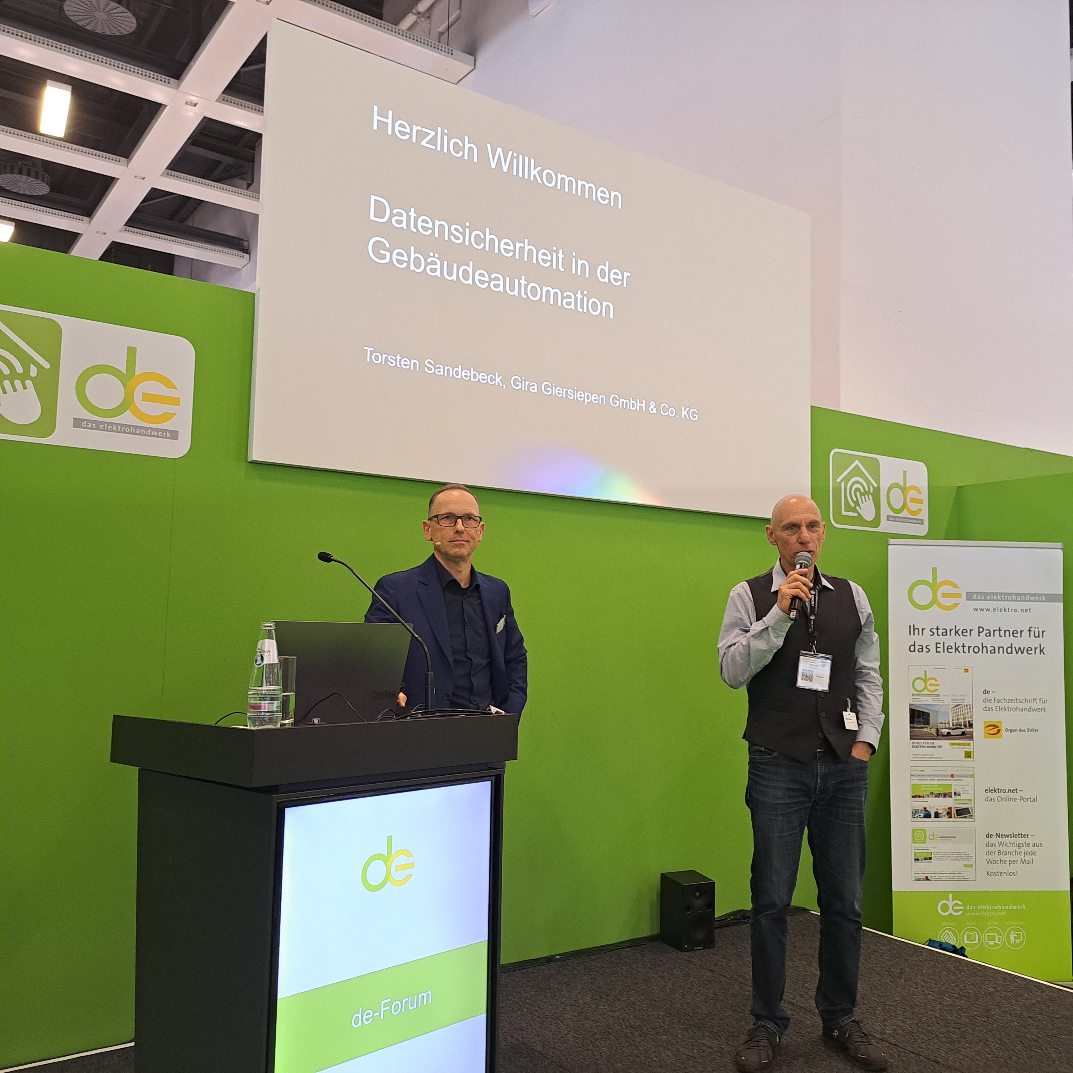 Blick ins de-Forum, Bühnensituation, Rednerpult und Leinwand mit zwei Referierenden auf der Bühne, grüne Wände mit de Logo